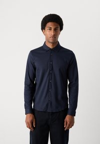 Marineblå langermet skjorte med knappedown-krage, med en glatt tekstur og subtil glans, kombinert med mørke jeans, båret av en modell.
