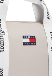 Μπεζ υφασμάτινη τσάντα ώμου με λευκό λουρί με λογότυπο, και ορθογώνιο κεντητό πατς που απεικονίζει το "TOMMY JEANS" σε κόκκινο, άσπρο και μπλε.