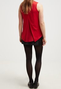 Blusa rossa senza maniche con dettaglio plissettato sulla schiena, indossata sopra una mini gonna nera con texture. Collant neri e scarpe completano l'outfit.