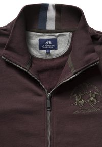Maroon zip-up jacket with gray inner lining, embroidered logo, and striped collar detailing. Features a metallic zipper and soft texture.