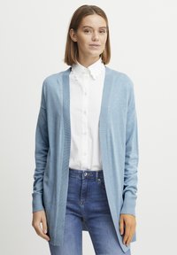 Svetlomodrý cardigan vyrobený z jemného materiálu, otvorený predný dizajn, rebrované manžety, dlhé rukávy, nosený cez bielu blúzku s volánmi a džínsy.