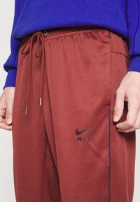 Maroon Nike joggers with elastic waistband and drawstring. Black "AIR" logo on front, side pocket visible, smooth synthetic fabric texture.