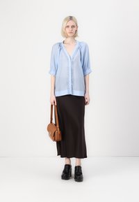 Lichtblauwe blouse met opgeblazen mouwen, decoratieve voorste afwerking; gecombineerd met een lange donkerbruine rok. Bruine schoudertas en zwarte platform schoenen.
