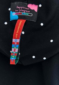 Czarna tkanina z małymi białymi kropkami, z kolorową metką z napisem "szczęście w trakcie realizacji" oraz zwrotami w jasnych kolorach.