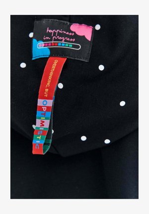 Čierna tkanina s malými bielymi bodkami, s farebnou etiketu, na ktorej je napísané "šťastie v procese" a frázy v jasných farbách.