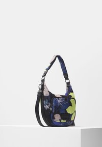 Torebka na ramię z regulowanym czarnym paskiem, wzór kwiatowy w kolorach niebieskim, zielonym i beżowym, przednia kieszeń zapinana na suwak oraz wyściełana rączka na białym tle.