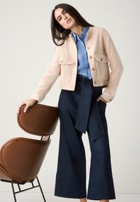 Hellbeige Cordjacke aus Cord mit Fronttaschen, kombiniert mit einem blauen Hemd und hoch geschnittenen, dunkelblauen, weiten Jeans. Brauner Stuhl im Hintergrund.