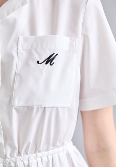 Hvit kortermet kjole med lomme som har svart broderi av "M." Innsmurt midje med glatt tekstur. Enkel, ren design.