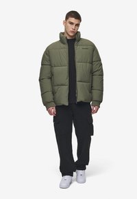 Jachetă pufoasă verde măsliniu cu guler înalt, având un design căptușit și manșete ribbate, purtată peste o cămașă neagră cu pantaloni cargo.