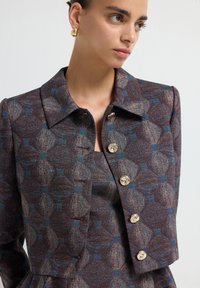 Chaqueta de tela texturizada en tonos marrón y azul con cuello, que presenta un patrón floral y botones dorados, con un corte entallado y hombros estructurados.