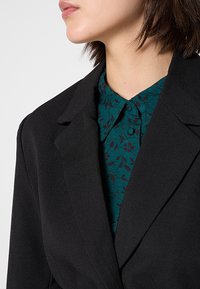 Blazer negro con solapas estructuradas, superpuesto a una camisa con patrón floral en tonos teal que presenta un cuello abotonado y una textura sutil.