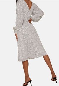 Silverfärgad paljettklänning med långa ärmar, V-formad rygg och figurnära midja. Längd till mitt på vaden med en svängig kjol. Svarta högklackade sandaler.