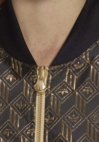 Černá bomber bunda s metalickým zlatým geometrickým vzorem, zlatý zip s úchytem ve tvaru mušle a žebrovaný límec. Texturovaný povrch s leskem.