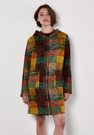 Donna che indossa un colorato cappotto con zip e cappuccio in patchwork, con tasche frontali, in piedi contro un muro bianco semplice.