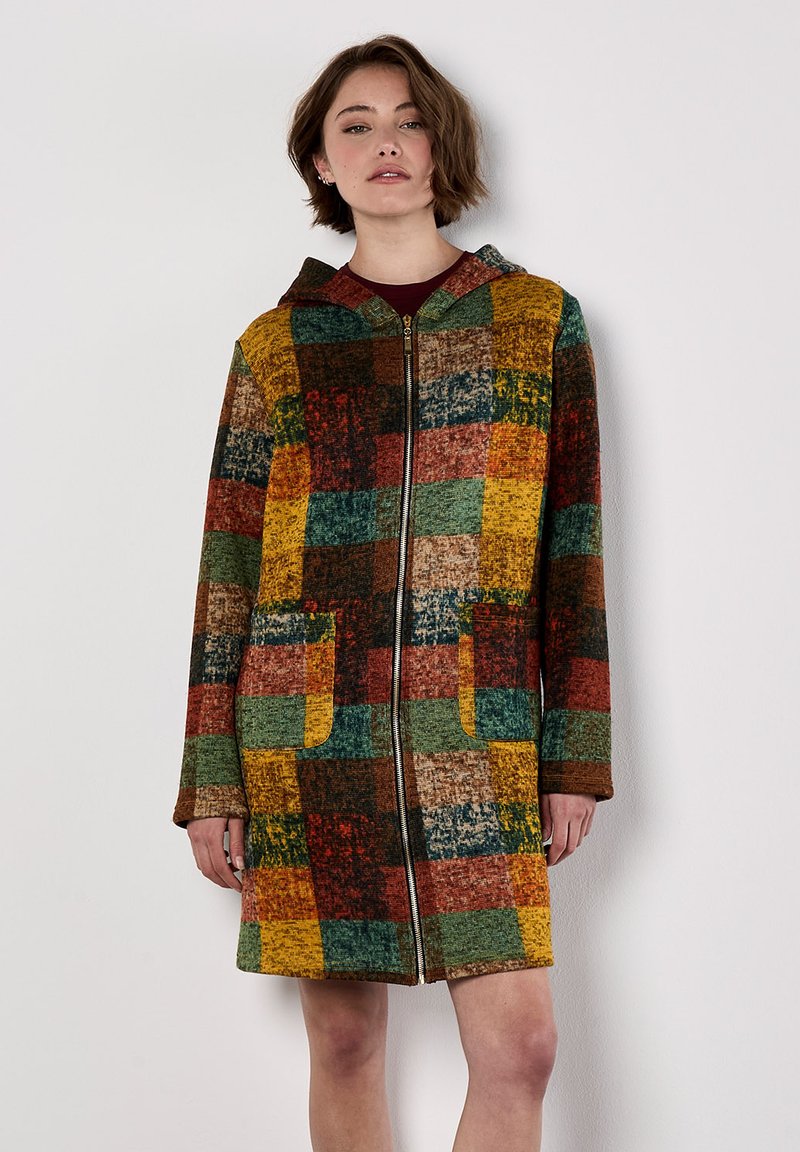 Vrouw in een kleurrijke patchwork ritsjack met capuchon en voorzakken, staande tegen een effen witte muur.