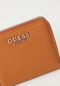 Braune, strukturierte Lederbörse mit silbernem Reißverschluss, der das eingeprägte „GUESS EST 1981“-Logo auf der Vorderseite zeigt. Kompakte rechteckige Form.