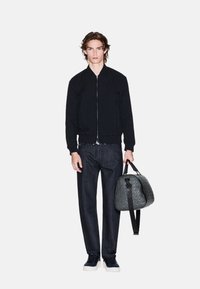 Giubbotto bomber nero in tessuto texturizzato, abbinato a pantaloni di jeans scuri e una borsa a tracolla nera a fantasia. Calzature casual incluse.
