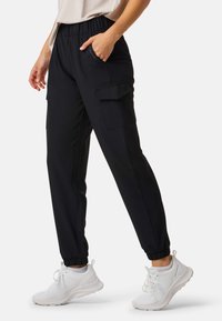 Person trägt schwarze Cargo-Jogger mit elastischen Bündchen und weiße Sportschuhe, steht mit einer Hand in der Tasche vor weißem Hintergrund.