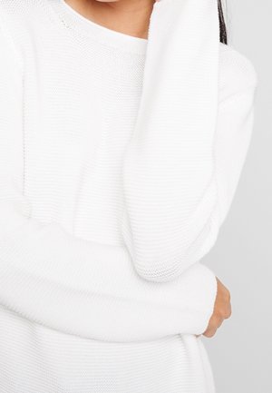 Frau trägt einen weißen gestrickten Pullover mit langen Ärmeln, eine Hand berührt ihren Hals und die andere ruht nahe ihrer Taille.