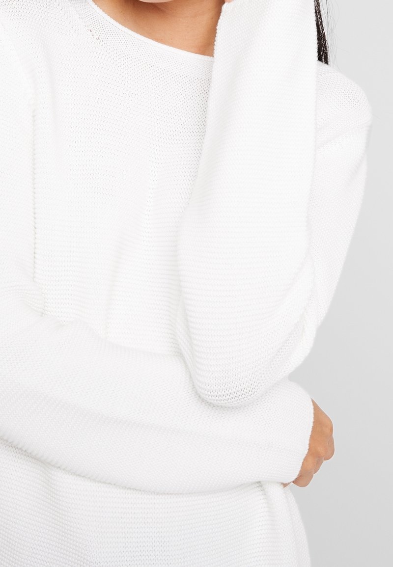 Femme portant un pull blanc en tricot à manches longues, une main touchant son cou et l'autre reposant près de sa taille.