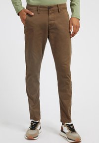 Pantalon en coton marron à coupe slim, avec deux poches latérales, une fermeture à bouton et des coutures contrastantes, porté avec des baskets beige et noires.