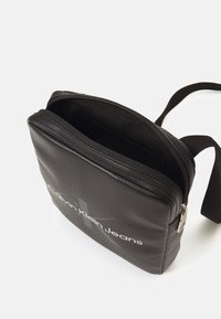 Borsa a tracolla in pelle nera con chiusura zip. Presenta un logo frontale bianco, una tracolla regolabile e una superficie liscia.