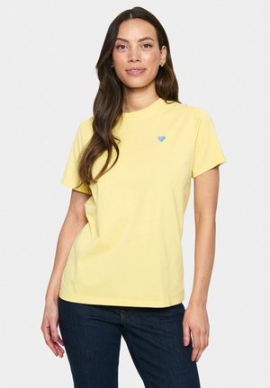 Gul bomulds T-shirt med korte ærmer, rund halsudskæring og lille blå hjertebroderi på brystet. Båret med mørke jeans.
