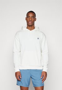 Hvita genser med hette, frontlomme, snor i hetta og en blå logo på brystet; kombinert med lyseblå shorts. Glatt tekstur.