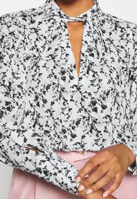 Blusa floreale bianca e nera con scollatura a chiave, maniche lunghe e tessuto strutturato. Posizionata sopra una vita rosa.
