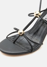 Μαύρο σανδάλι με λεπτές λωρίδες, χρυσές στρογγυλές διακοσμήσεις και λεπτομέρεια υφή πέλματος, τοποθετημένο σε λευκό φόντο.