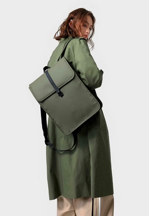 Grüner Stoffrucksack mit Klappenverschluss, schwarzem Tragegurt und schlanker, rechteckiger Form. Wird auf dem Rücken getragen und kombiniert mit einem langen Mantel und hellen Hosen.