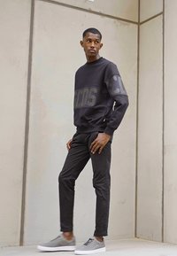 Fekete pulóver nagy szürke felirattal az ujjakon, slim-fit sötét nadrággal és szürke sportcipővel párosítva. Sima szövet minimalista dizájnnal.