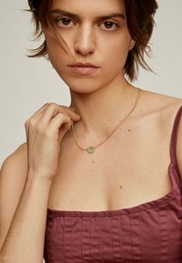 Collar de cadena de oro con un pequeño colgante redondo verde. El fondo es neutro, lo que realza los detalles del collar.