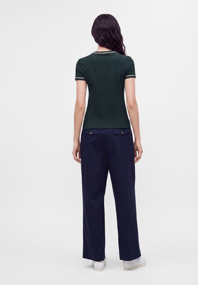 Femme aux longs cheveux foncés, vue de dos, portant un t-shirt vert foncé à manches courtes et un pantalon large bleu marine avec des baskets blanches.