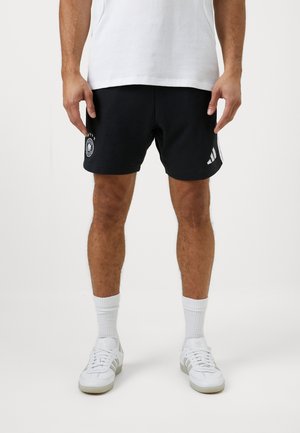 Juodi sportiniai šortai su siuvinėtu logotipu ir trimis baltais dryžiais šonuose, pagaminti iš švelnios medžiagos, derinami su baltais sportiniais bateliais ir kojinėmis.