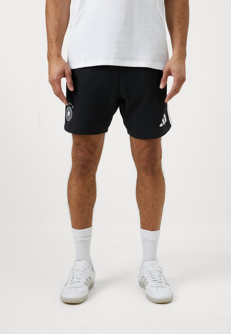 Μαύρες αθλητικές βερμούδες με κεντητό λογότυπο και τρεις λευκές ρίγες στις πλευρές, φτιαγμένες από μαλακό ύφασμα, συνδυασμένες με λευκά αθλητικά παπούτσια και κάλτσες.