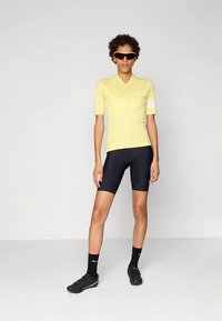 Geel fietsshirt met korte mouwen en een ritssluiting aan de voorkant, gecombineerd met zwarte fietsshorts. Model draagt getinte zonnebril en zwarte fietsschoenen.