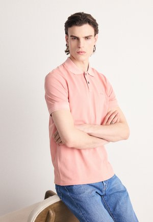 Camiseta polo rosa claro con cuello, tapeta de dos botones y mangas cortas, combinada con jeans de denim azul. Diseño simple, textura suave.