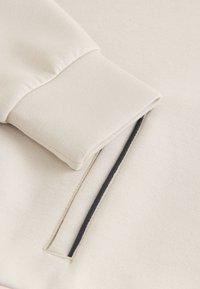 Manche de sweat-shirt couleur crème avec un poignet côtelé ; présente une fente sur la couture latérale avec un bord noir contrastant. Tissu doux et texturé.
