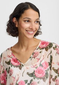 Blumenbluse mit einem V-Ausschnitt, aus leichtem, transparentem Stoff. Sie zeigt pinke Rosen und grüne Blätter auf einem hellen Hintergrund.