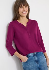 Femme souriante appuyée contre un mur blanc, portant une chemise violette à manches longues avec trois boutons et un jean bleu, une main dans la poche.