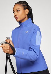Svijetloplavi softshell jakna s visokim ovratnikom, džepom na patentni zatvarač na prsima i teksturiranim materijalom; opremljena za vanjske aktivnosti, sa podešivim manžetama.