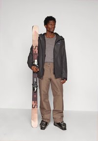 Equipamento de esqui composto por um casaco impermeável preto, camisa cinza, calças bege, botas de neve pretas e um esqui com design rosa e castanho.