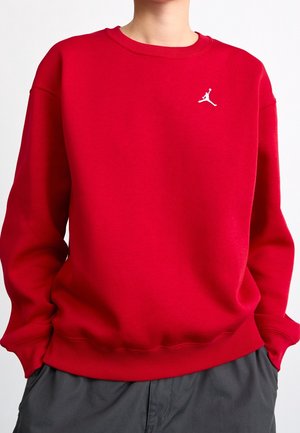 Personne portant un sweat-shirt rouge à col rond avec un petit logo blanc Jumpman sur le côté gauche de la poitrine et un pantalon gris foncé.