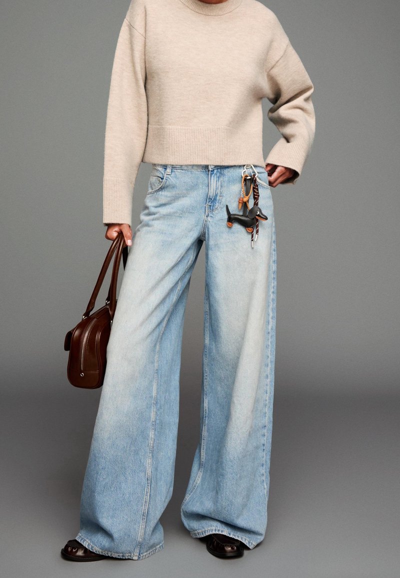 Personne portant un pull court beige, un jean large bleu clair avec un porte-clés chien noir, tenant un sac à main marron sur fond gris.