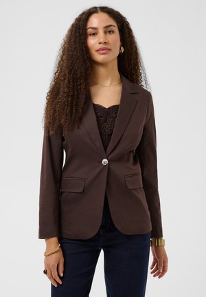 Femme aux longs cheveux bouclés portant un blazer marron foncé à un seul bouton sur un top en dentelle noire et un jean foncé sur un fond uni.