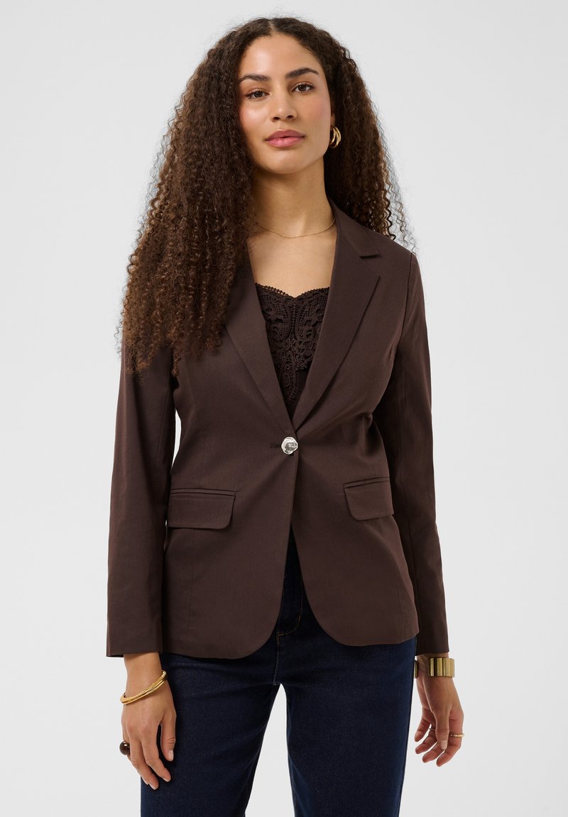 Femme aux longs cheveux bouclés portant un blazer marron foncé à un seul bouton sur un top en dentelle noire et un jean foncé sur un fond uni.