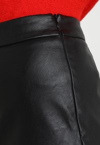 Schwarzer Kunstlederrock mit glatter Textur, hoher Taille und einem verdeckten Reißverschluss auf der Rückseite, vor einem roten Hintergrund festgehalten.