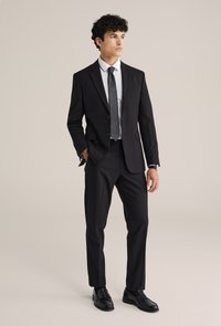 Costume noir à deux boutons, associé à une chemise blanche et une cravate grise. Un pantalon ajusté et des chaussures noires brillantes complètent le look.