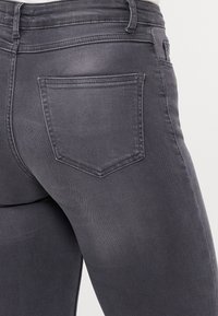 Vue arrière d'un jean en denim gris porté, montrant la poche arrière droite et la ceinture avec passants contre un fond uni.